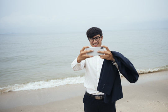穿着白衬衫的黑发男人站在沙滩上看不清天空，用智能手机自拍。