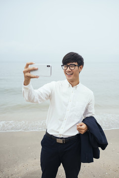 穿着白衬衫的黑发男人站在沙滩上看不清天空，用智能手机自拍，垂直。