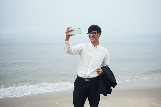 黑发白衣人站在阴沉的沙滩上，用智能手机自拍。