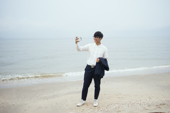 黑发白衣人站在阴沉的沙滩上，用智能手机自拍，全拍。