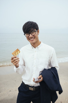 黑发小伙穿着白衬衫站在沙滩上，阴沉的天空中，出示信用卡，垂直。