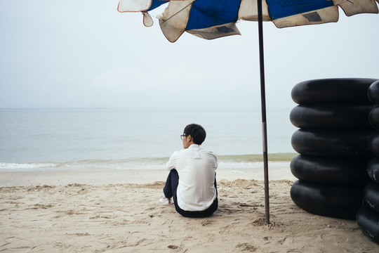 黑发小伙穿着白衬衫背对着镜头坐在沙滩上的黑色游泳圈旁，环顾大海。