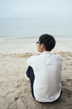 穿着白衬衫的黑发男人坐在沙滩上对着镜头环顾大海。