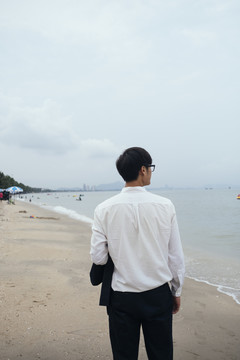 穿着白衬衫的黑发男人从沙滩上的摄像机里走出来，看着大海。