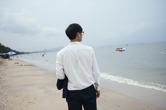 黑发小伙穿着白衬衫走出相机在沙滩上望着大海，水平。