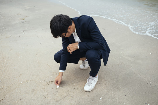 黑发男穿着深色西装坐在沙滩上捡贝壳。