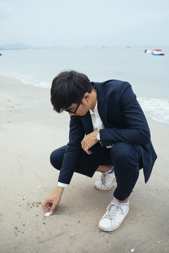 黑头发的家伙穿着深色西装坐在沙滩上拿起贝壳，垂直。