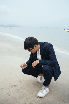黑头发的家伙穿着深色西装坐在沙滩上看着他手里的贝壳。