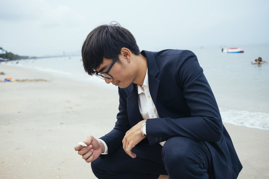 黑头发的家伙穿着深色西装坐在沙滩上看着手里的贝壳，水平。