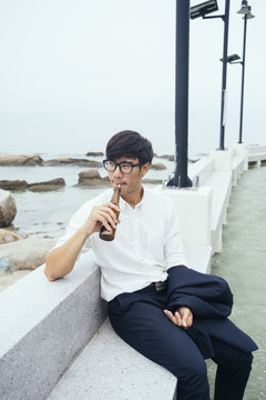 黑发汉子穿着白衬衫，坐在大理石长凳上，沿着码头边满是岩石的海面，用吸管喝着饮料。