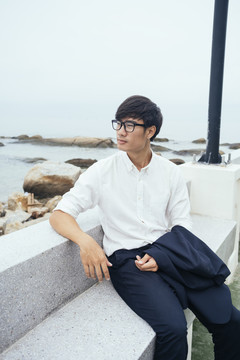 黑发汉子穿着白衬衫坐在大理石长凳上，沿着码头的海边布满岩石，笔直。