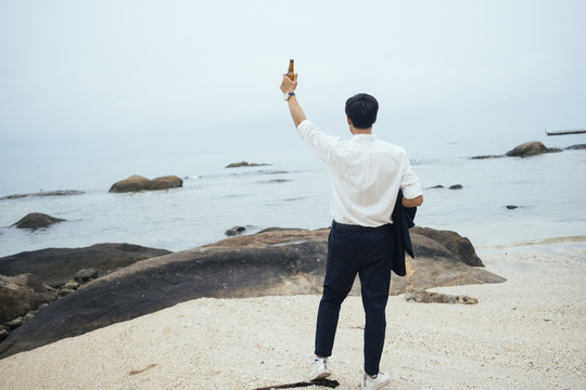 黑发汉子穿着白衬衫站在海边的沙滩上，他举起手中的酒瓶向天空敬酒。