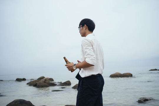 在阴沉的日子里，一个穿着白衬衫的黑发男人手里拿着瓶子站在海边的低角度镜头。