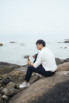 黑发汉子穿着白衬衫坐在海边的岩石上，手里拿着瓶子，想着自己的成功。