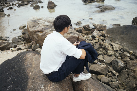 穿着白衬衫的黑发男人坐在海边的岩石上，手里拿着瓶子，看着大海，想着自己的失败。