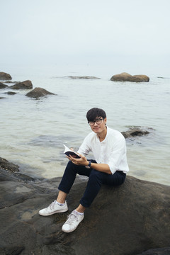 黑发小伙穿着白衬衫坐在海边的岩石上看书，看着相机。