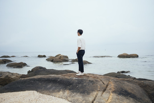 内省的黑发人穿着白衬衫站在海边的岩石上。