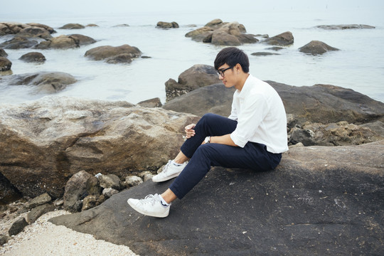 黑发男穿着白衬衫坐在海边的岩石上，抱着膝盖。