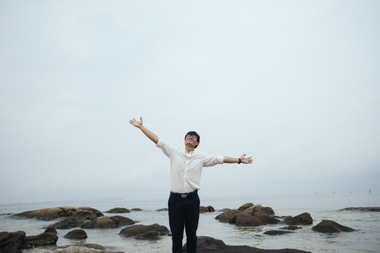 黑发白衣人站在海边的岩石上，双臂搭在头上，像是要拥抱天空。