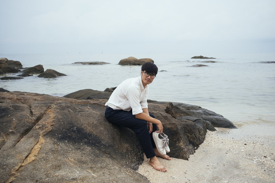 黑发白衣人脱下鞋子，坐在海边的岩石上。
