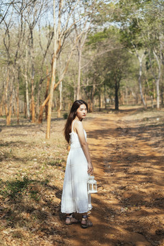 美丽的年轻女子穿着优雅的白色礼服，在秋林里提着白色的油灯。