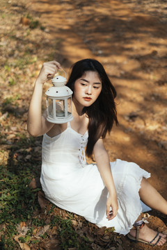 美丽的年轻女子身穿优雅的白色礼服，手持古董灯笼，坐在泥土上。