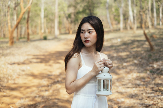 美丽的年轻女子穿着优雅的白色礼服，双手捧着古董油灯。在森林里迷路的女孩。