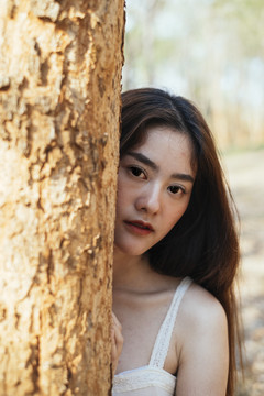 特写镜头：一位身穿优雅白色连衣裙的年轻漂亮女子站在一棵树后。
