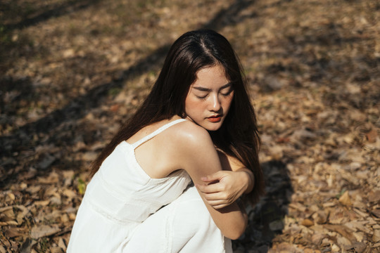 亚洲美丽的年轻女子穿着优雅的白裙坐在秋林里。在森林里迷路的女孩。