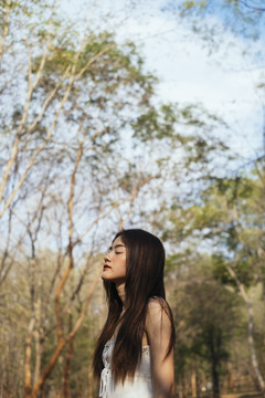 美丽的年轻女子穿着优雅的白裙，闭着眼睛站在森林里。