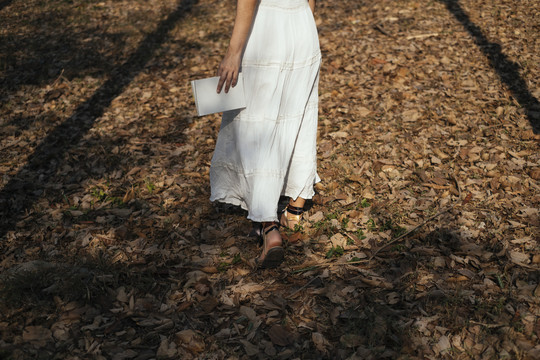 一个穿着白衣服的女人拿着一本书独自在森林里走着。