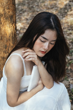 秋林里，一位穿着优雅白裙的美丽年轻女子抱着一本书的特写镜头。