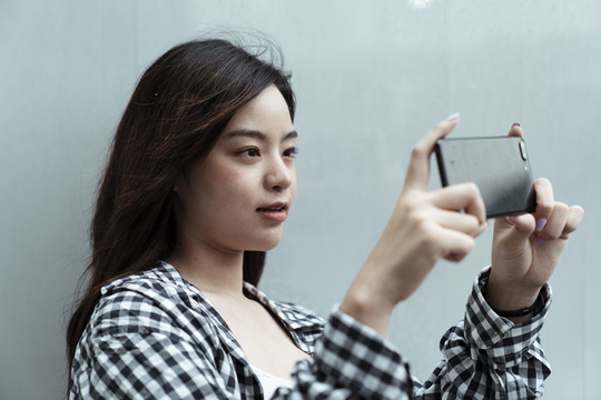 黑格纹长发女孩站在墙前，举着智能手机拍照。