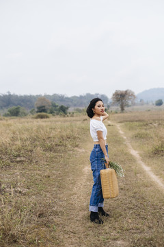 身穿白衬衫和蓝色牛仔裤的亚洲女孩的侧视图，她提着篮子准备在郊野野餐。