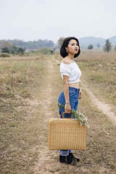 身穿白衬衫和蓝色牛仔裤的亚洲女孩的侧视图，她提着篮子准备在郊野野餐。