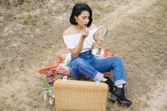 年轻性感的亚洲女孩正在野餐，坐在垫子上，化妆，拿着一个便携式镜子，享受化妆的乐趣。