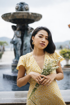 年轻的亚洲女人穿着黄色的裙子，手持白色的花束站在室外的喷泉旁。