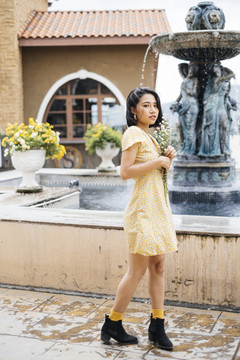 身着黄色连衣裙的亚洲年轻女子，手持白色花束站在室外喷泉旁。