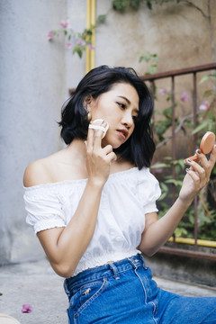 年轻性感的亚洲女人穿着白衬衫和蓝色牛仔裤坐在楼梯上走出家门，化着妆，看着一面小镜子。