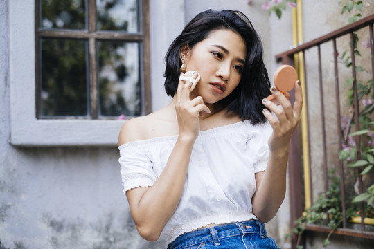 年轻性感的亚洲女人穿着白衬衫和蓝色牛仔裤坐在楼梯上走出家门，化着妆，看着一面小镜子。