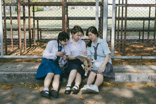 三个穿着不同制服的漂亮亚泰学生坐在人行道上，互相帮助，一起做作业。