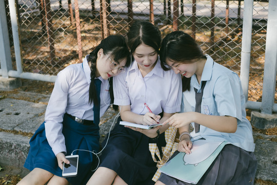 三个穿着不同制服的漂亮亚泰学生坐在人行道上，互相帮助，一起做作业。