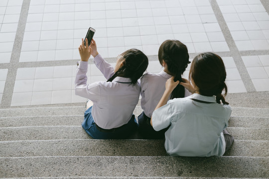 三个穿着不同制服的美丽亚泰学生坐在楼梯上用智能手机拍照或自拍。社交生活。在学校上网。