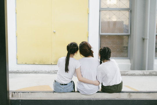 后视图-三个美丽的亚泰学生穿着白色休闲衬衫坐在阳台上看建筑。