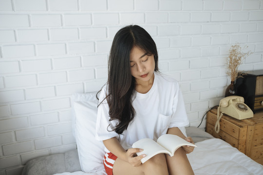 可爱的亚洲年轻女子坐在床上，把一本书放在膝盖上看书。
