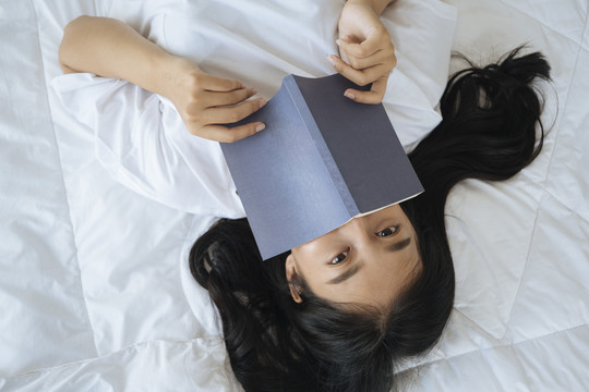 亚洲年轻女子睡在床上，脸上盖着书的俯视图。