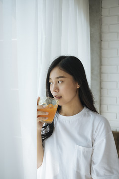 美丽的亚洲年轻女子手持装满橙汁的玻璃杯作为早餐，背景是白色的窗帘。