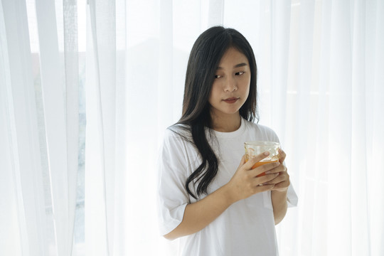 美丽的亚洲年轻女子手持装满橙汁的玻璃杯作为早餐，背景是白色的窗帘。