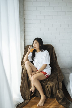 年轻的亚洲女士在卧室的皮沙发上喝着一杯橙汁。