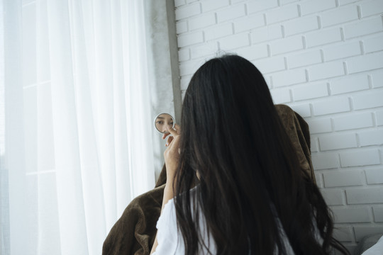 年轻的亚洲女人拿着一面小镜子，在卧室里化妆。后视图。